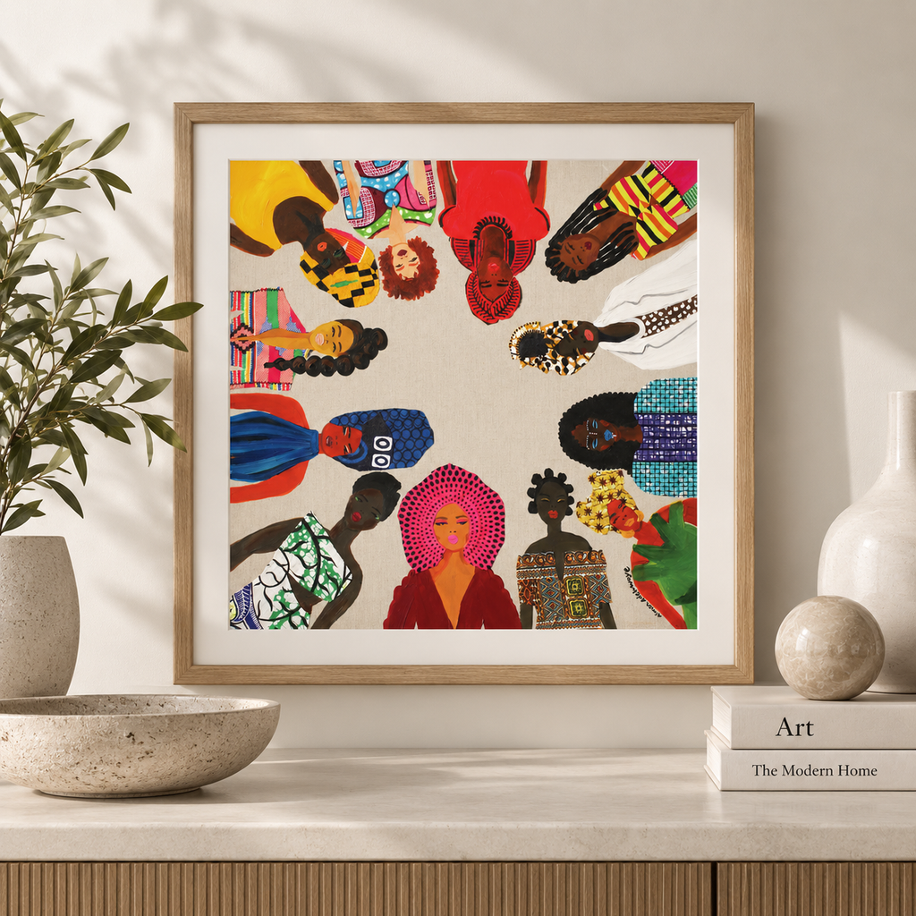 A framed art print featuring a colorful circular collage of stylized women, displayed on a light marble console table with decorative plants, ceramic vases, and a book in a styled interior setting.