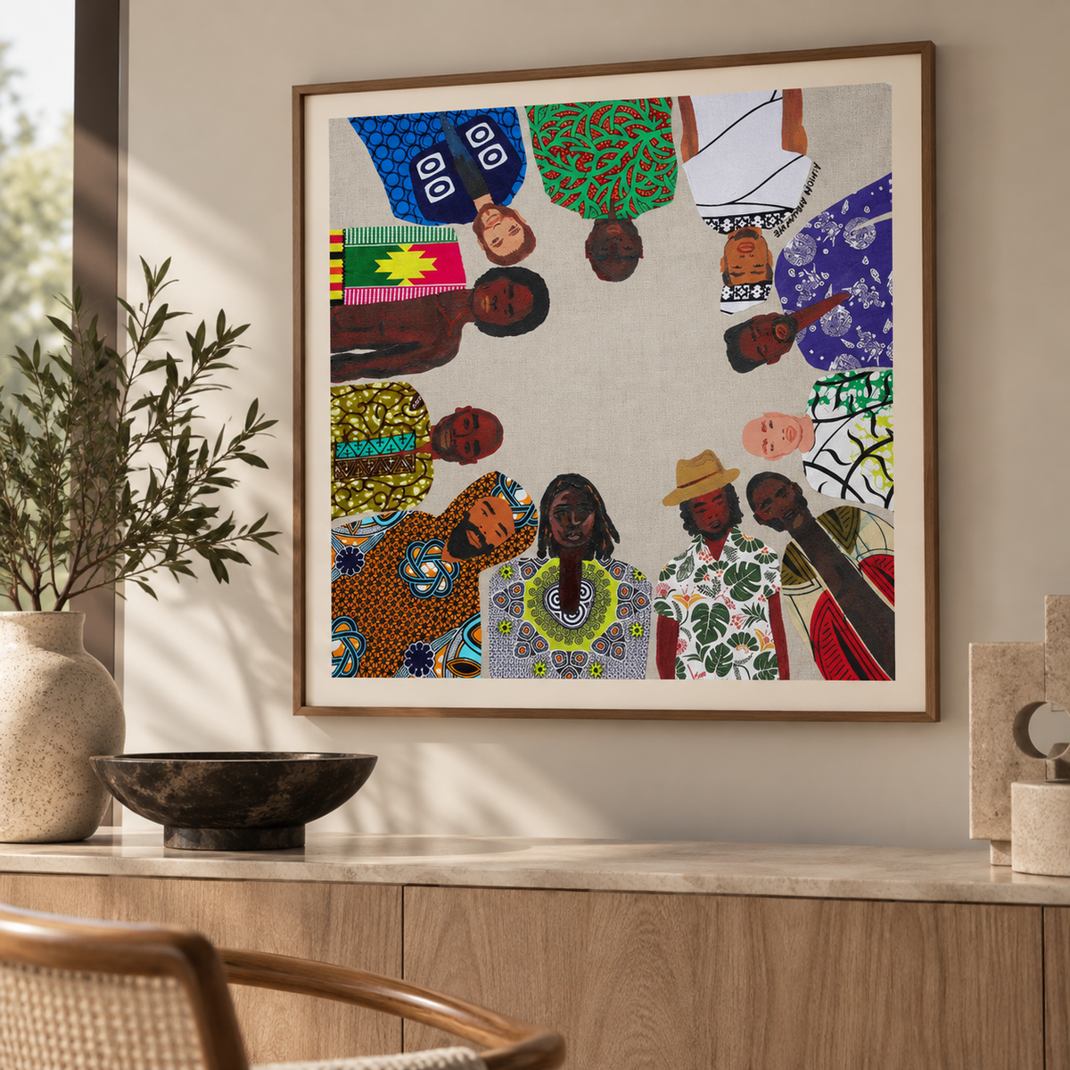 A framed art print featuring a circular collage of diverse men in colorful African patterned clothing, displayed on a light beige wall above a wooden console table with decorative items.