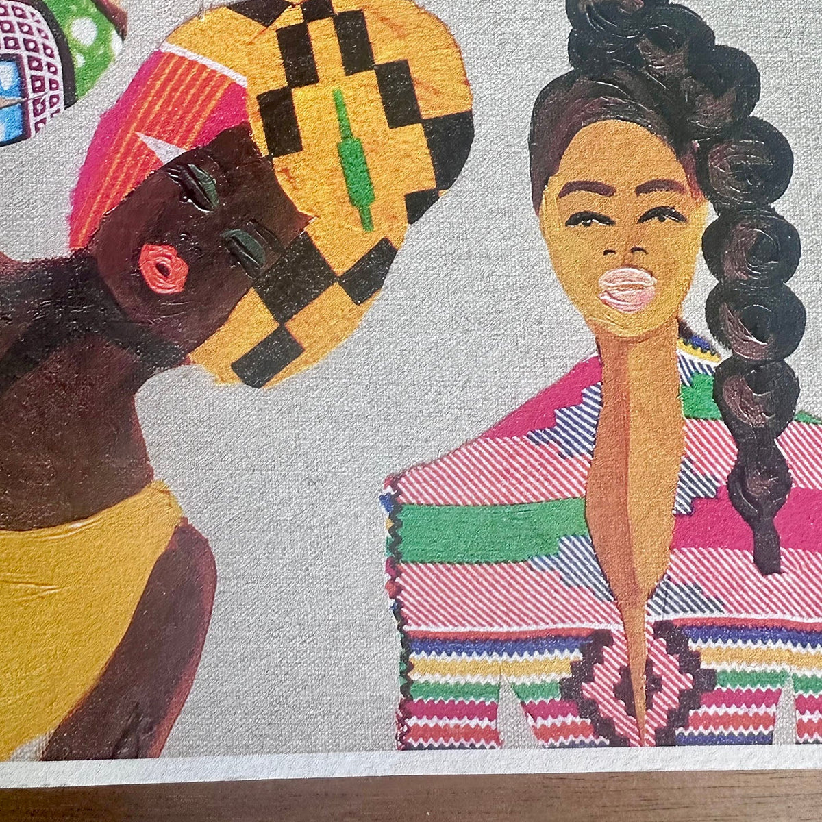 A close-up of a colorful print artwork showing stylized  Black women with bold patterns and warm skin tones on a light textured background. A yellow head-tie, natural afro hair, a high ponytail, and bold colourful suit. 