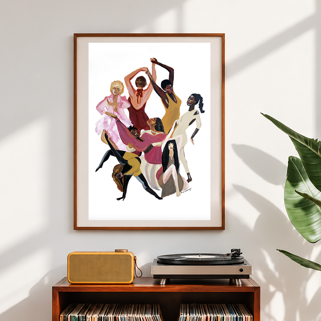 A framed art print featuring a stylized illustration of eight women dancing together, displayed on a white wall above a wooden console table with a record player and vinyl records. A green plant leaf is visible on the right side.